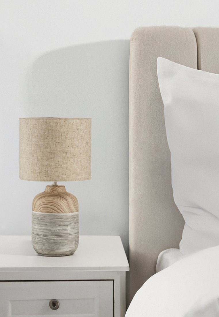 Tischlampe mit einem runden Holzfuß, der ein helles Holzmaserungsmuster aufweist, gekrönt von einem beigefarbenen Stoffschirm. Neben einem hellen gepolsterten Kopfteil platziert.