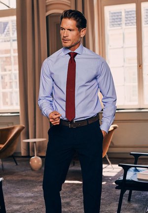 Lichtblauw gestreepte dress shirt met een rode stropdas, donkere broek en een leren riem in bruin. De setting heeft zachte verlichting en minimalistisch meubilair.