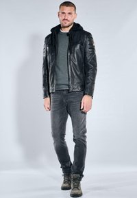 Mann steht mit entspannten Händen, trägt schwarze Lederjacke mit Kapuze, grauen Pullover, ausgewaschene schwarze Jeans und graue Schnürstiefel.