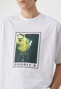 Valge puuvillane t-särk, millel on rohelises ja mustas värvitoonis ristkülikukujuline graafiline print, mille all on tekst "DOUBLE A". Lühikesed varrukad.