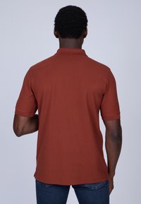 Polo à manches courtes en orange rouille, fabriqué en coton. Il présente un col côtelé et un ourlet droit, avec des fentes latérales en bas.