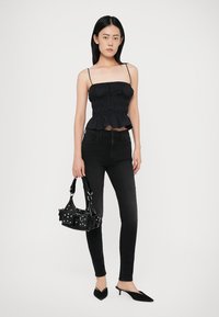 Top preto sem alças com textura franzida, combinado com jeans skinny pretos. A modelo segura uma pequena bolsa preta com tachas prateadas e usa sapatos planos pretos de bico.