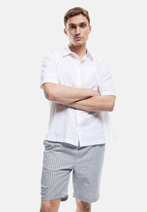 Camisa de lino blanca de manga corta con un bolsillo, combinada con pantalones cortos a rayas en azul y blanco, ambos con un corte relajado y tejido transpirable.