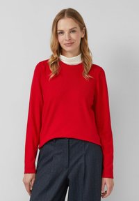 Czerwona dzianinowa koszulka z długim rękawem i okrągłym dekoltem, noszona na białym swetrze z golfem, w zestawie z ciemnymi spodniami w paski. Gładka faktura.