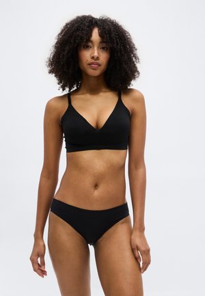 Γυναίκα με σγουρά μαλλιά που φοράει μαύρο σουτιέν και μαύρο εσώρουχο, στέκεται μπροστά σε απλό λευκό φόντο.