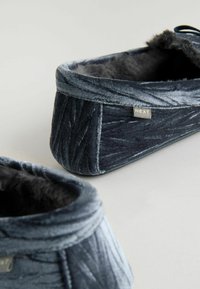 Kék bársonybéli papucsok, műszőrme béléssel, lekerekített formával és texturált felülettel, a "NEXT" felirattal ellátott kis címkével.