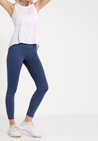 Débardeur blanc léger à coupe ample, associé à des leggings bleu foncé ajustés et des chaussures de sport blanches avec des détails en mesh.