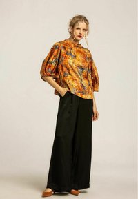 Färgglad, mönstrad puffärmad blus i slätt tyg, tillsammans med vida svarta byxor och bruna skor mot en neutral bakgrund.