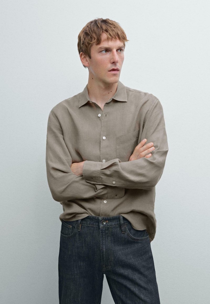 Beige linen button-up shirt with a chest pocket, paired with dark blue denim jeans. Features long sleeves and a relaxed fit.