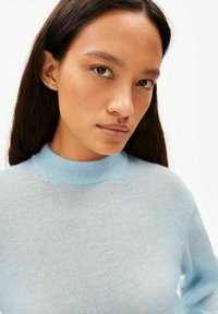 Hellblauer Strickpullover mit hohem Kragen, weicher Textur und dezentem Muster. Nahaufnahme des Oberkörpers, die den Kragen zeigt.