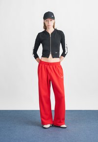 Crna zip-up kratak jakna s bijelim bočnim prugama i prednjim džepom, uparena s labavim crvenim trenirkama koje imaju bijeli logo. Crna kapa.