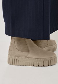 Beige chunky platform boots with elastic side panels and textured soles, paired with navy pinstripe trousers. Smooth suede finish.