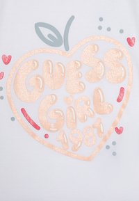 Hvit skjorte med stor glitrende ferskenfarget tekst i hjerteform, som sier "GUESS GIRL 1981," med små røde hjerter og et grønt blad som aksent.