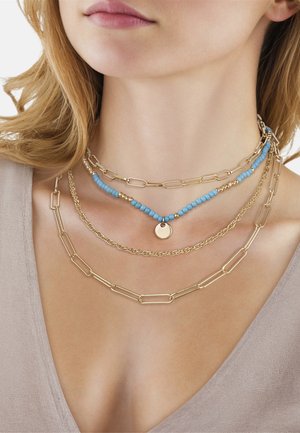 Collares de cadenas doradas en capas con cuentas azules y doradas y un colgante redondo de oro, usados en el cuello de una mujer con una blusa beige.