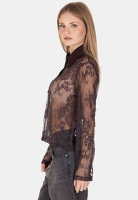 Giovane donna che indossa una blusa nera in pizzo floreale trasparente con maniche lunghe e jeans scuri, in piedi di profilo contro uno sfondo semplice.