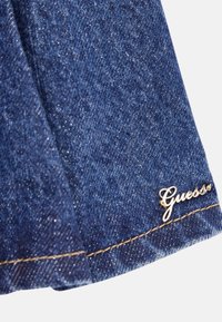 Denimstof i mørkeblå med en subtil glans; har et guld "Guess" logo-tag og kontrasterende beige syninger langs kanten.