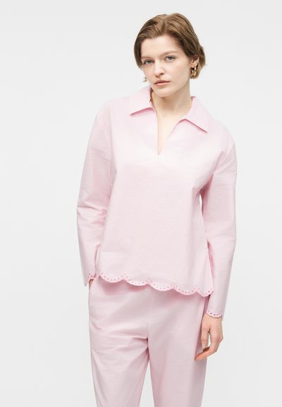 Νεαρή γυναίκα με κοντά μαλλιά που φοράει ροζ ανοιχτό μακρυμάνικο πουκάμισο με κυματιστή λεπτομέρεια στο τελείωμα και παντελόνι στο ίδιο χρώμα, στέκεται μπροστά σε λευκό φόντο.