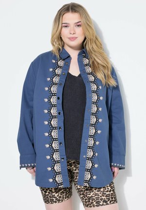 Camicia azzurra con bottoni decorata con intricati ricami beige sul davanti, con una maglietta nera testurizzata sotto e pantaloni corti leopardati.