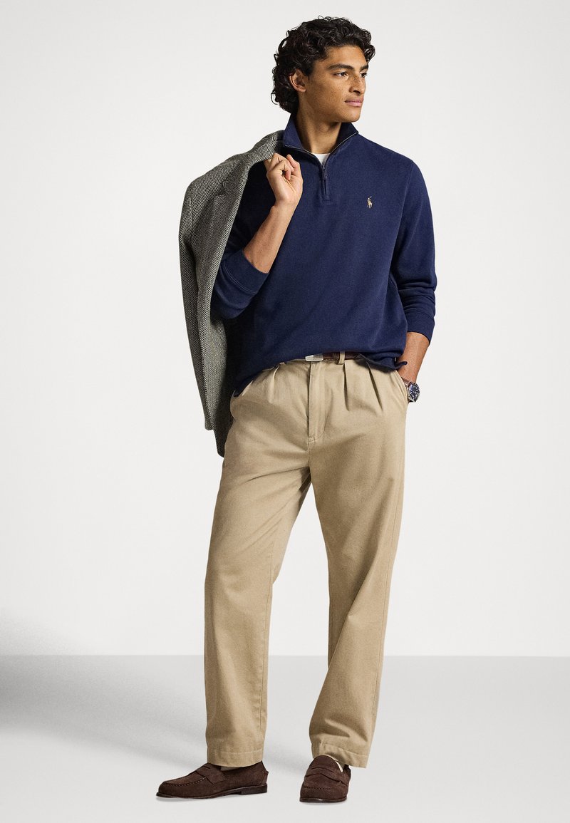 Man in navy quarter-zip sweater and khaki pants holds gray coat over shoulder, wearing brown loafers and wristwatch, looking right.