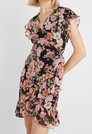 Kobieta w czarnej sukience kopertowej z różowym, białym i pomarańczowym kwiatowym wzorem, z falbanami na krótkich rękawach i u dołu sukienki.