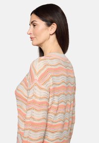 Pull en tricot présentant un motif ondulé dans des tons crème, rose et orange avec une texture à mailles ajourées, offrant respirabilité et style superposé.
