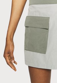 Ljusgrå kjol med en stor grön tygficka. Materialet verkar lätt och har en slät textur. Synliga sömdetaljer lyfter fram designen.