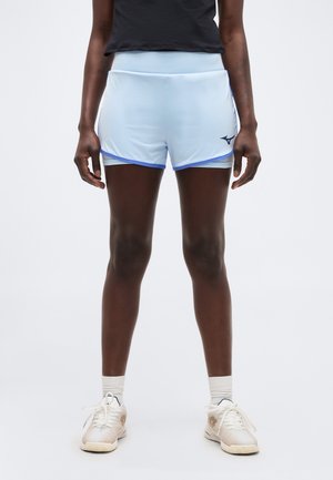 Sportoló világoskék sportnadrágban sötétkék szegéllyel, fehér zokniban és bézs színű futócipőben, egyszerű fehér háttér előtt.
