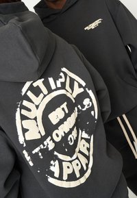 Des sweats à capuche noirs avec des impressions graphiques blanches au dos et sur la poitrine, présentant un design rond avec des lettres vieillies et une texture douce.