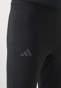 Czarne legginsy sportowe o gładkiej fakturze, z subtelnym matowym logo w postaci trzech pasków w szarym kolorze na lewej udzie oraz płaskimi szwami.