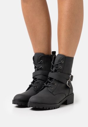 Botas negras hasta el tobillo hechas de material sintético suave, con cordones en la parte delantera, correa con hebilla y suela gruesa texturizada.