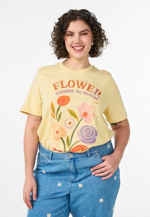 Smilende kvinde med kort krøllet hår iført en gul blomster T-shirt med teksten "FLOWER CHOOSE TO BLOOM" og blå jeans med små hvide blomster.