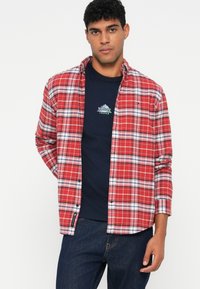 Uomo che indossa una camicia a quadri rossa e bianca sopra una maglietta navy di Tommy Jeans e jeans blu scuro, in piedi contro uno sfondo bianco.