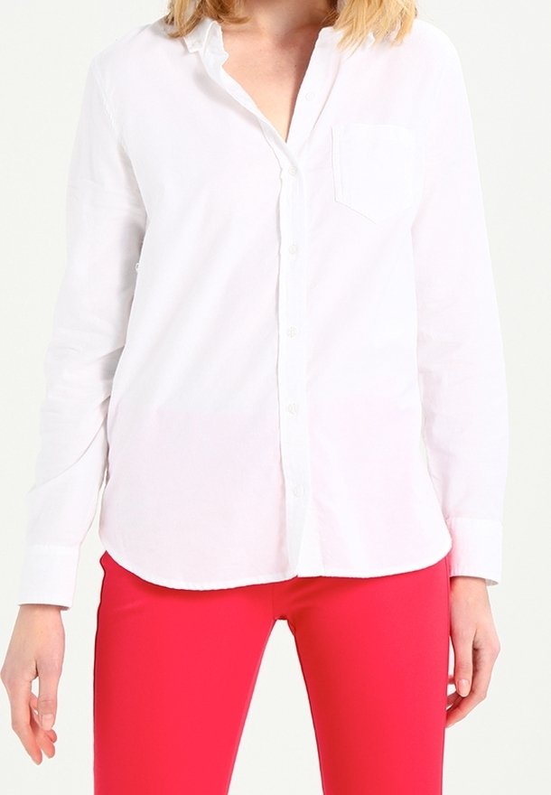 Femme portant une chemise blanche à manches longues boutonnée et un pantalon rouge vif, debout devant un fond clair uni.