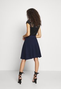 Top preto de renda com mangas curtas e padrões florais, combinado com uma saia rodada azul-marinho. Apresenta uma cintura ajustada e uma textura suave.
