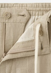 Beżowe lniane spodnie z elastycznym pasem, zamkiem błyskawicznym, zapięciem na guzik oraz białym sznurkiem częściowo rozwiązanym, ukazującym wewnętrzną podszewkę.