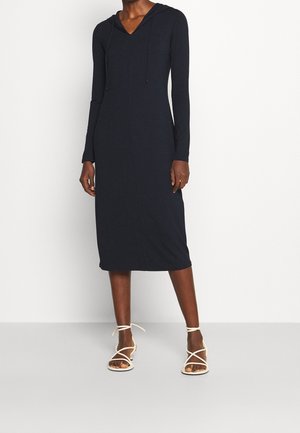 Kobieta ubrana w czarną, długorękawową, midi sukienkę z kapturem, w połączeniu z białymi sandałkami na wysokim obcasie z paskami, stojąca na tle gładkiej białej ściany.