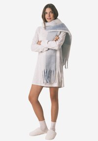 Lett blå og hvid ombre tørklæde med frynseender, båret over en kremfarvet kjole, parret med hvide teksturerede slippers. Glat stoftekstur.