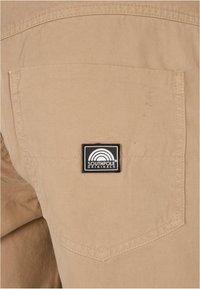 Beige kangas, jossa on sileä pinta; sisältää takataskun ja mustan neliön muotoisen tarran, jossa on valkoista tekstiä ja sateenkaari grafiikka.