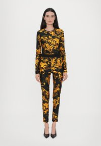 Abito in tessuto elasticizzato nero con audaci motivi floreali gialli e dorati, dotato di maniche lunghe e pantaloni aderenti, abbinato a tacchi neri.