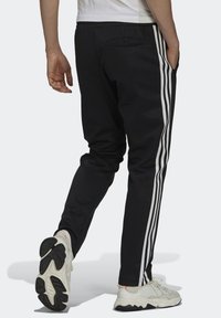 Černé joggingové kalhoty s bílými bočními pruhy, elastickým pasem a zadní kapsou, v kombinaci se světle barevnými sportovními botami.