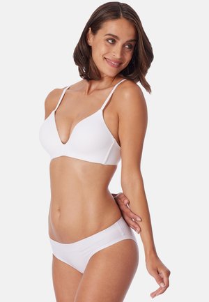 Femme aux cheveux bruns mi-longs portant un soutien-gorge blanc et une culotte assortie, souriante et regardant vers sa gauche.