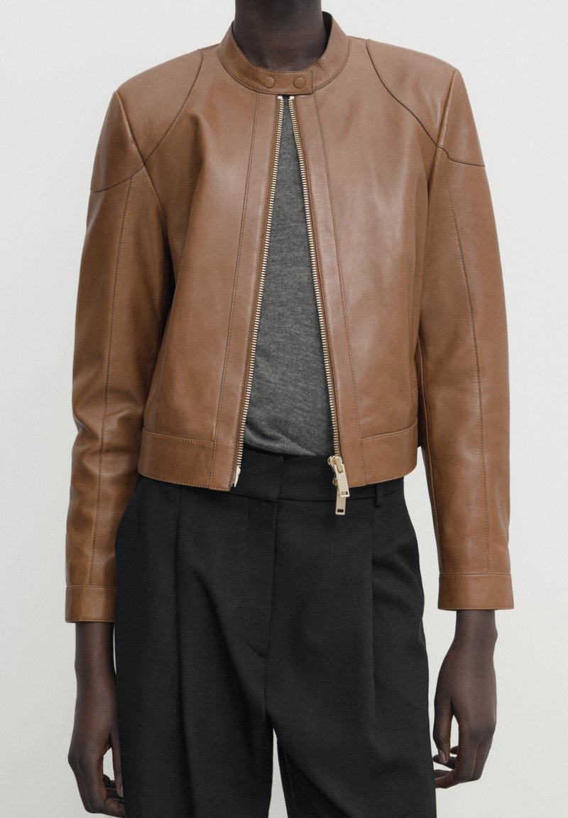 Mörkhyad person som bär en kort klipp brun läderjacka med dragkedja, grå tröja och svarta högmidjade byxor mot en enfärgad bakgrund.