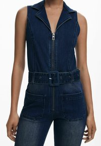 Denim overal má predný zips, golier, opasok v páse a dve predné vrecká. Tmavomodrá farba s vyblednutou textúrou a štruktúrovaným dizajnom.