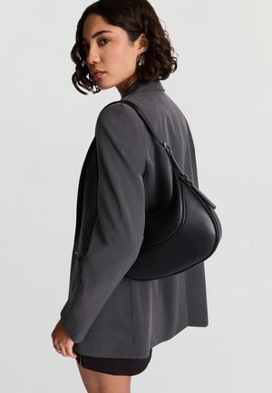 Donna con capelli corti e ricci che indossa un blazer grigio e una gonna nera, con una borsa a tracolla nera a forma di mezzaluna.