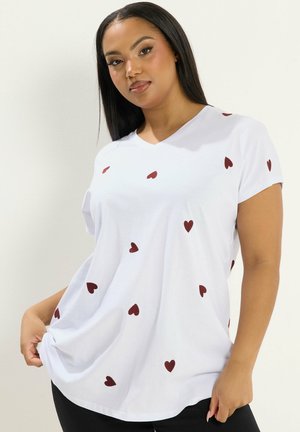 Kvinde med langt sort hår iført en hvid V-hals T-shirt med spredte røde hjerteprint, der holder i kanten af skjorten på den ene side.