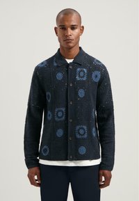 Cardigan tricotat de culoare albastru închis, cu modele florale albastre, guler și închidere cu nasturi. Are o țesătură texturată și mâneci lungi.