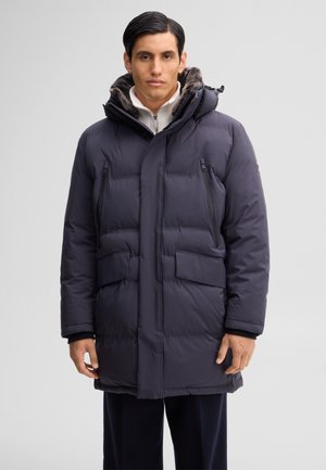 Mann trägt eine dunkel gepolsterte Winterjacke mit pelzgefütterter Kapuze und Reißverschlusstaschen, steht vor einem einfarbigen hellgrauen Hintergrund.