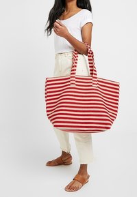 Grand sac fourre-tout rayé rouge et blanc avec de larges poignées en tissu, fabriqué en un matériau résistant. Le sac a un haut ouvert et une forme rectangulaire.