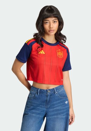 Žena s dugom tamnom kosom koja nosi crvenu i tamnoplavu kratku španjolsku nogometnu majicu te plave Adidas traperice, stoji na svijetloj pozadini.