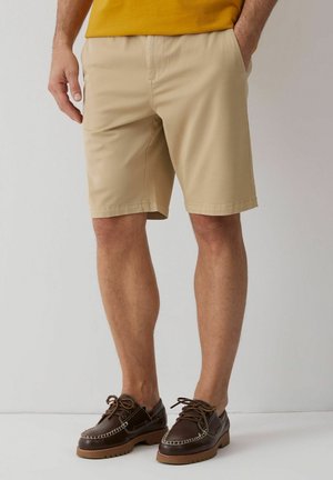 Mężczyzna w beżowych szortach i ciemnobrązowych sznurowanych butach żeglarskich, stojący na jednolitym jasnym tle.